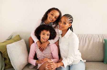 Téléchargez les photos : Le bonheur familial. afro-américaine mère et filles embrassant posant ensemble souriant à la caméra assis sur canapé dans le salon moderne à la maison. Concept de maternité heureuse - en image libre de droit