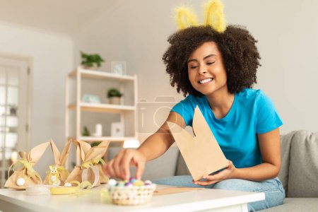 Téléchargez les photos : Portrait d'une belle femme afro-américaine préparant des cadeaux de Pâques à la maison, heureuse jeune femme noire souriante portant des oreilles de lapin bandeau d'emballage friandises festives dans des sacs en papier artisanal - en image libre de droit