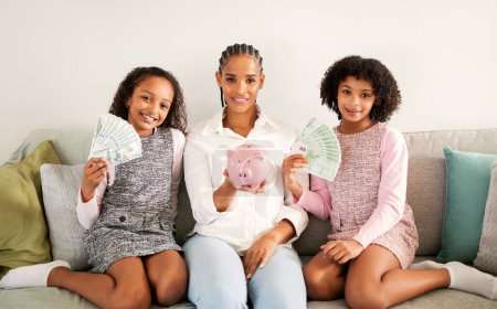 Photo for Financial Offer. African American Mother And Daughters Holding Piggybank With Family Savings Sitting On Sofa Smiling To Camera Posing At Home. Bank Service And Finance Safety - Royalty Free Image