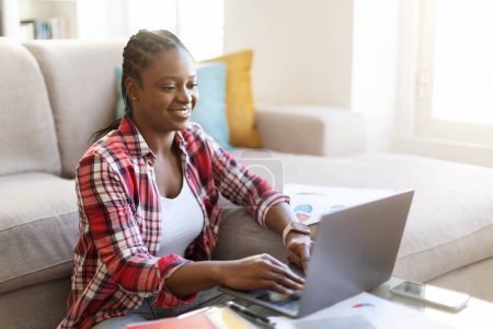 Photo for Positive pretty millennial black woman in casual outfit sitting on floor in living room, typing on laptop keyboard and smiling, female freelancer working from home, copy space. Remote job - Royalty Free Image