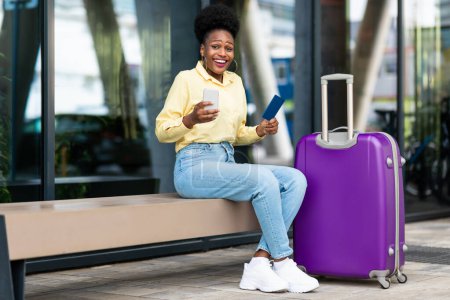Téléchargez les photos : Lady touristique afro-américaine excitée utilisant la réservation de téléphone portable billets de voyage en application sourire à la caméra assis avec valise à l'extérieur près du terminal de l'aéroport. Vacances, Enfin - en image libre de droit