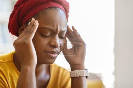 Photo for Exhausting migraine concept. Unhappy young black woman wearing red african turban suffering from headache at home, sitting on couch with closed eyes, touching temples, closeup, copy space - Royalty Free Image