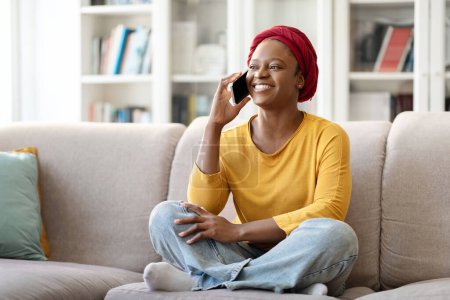 Foto de Feliz alegre joven negra en casual vistiendo turbante africano rojo sentado en el sofá en casa, hablando por teléfono con un amigo o amante, compartiendo noticias, mirando el espacio de copia y sonriendo - Imagen libre de derechos