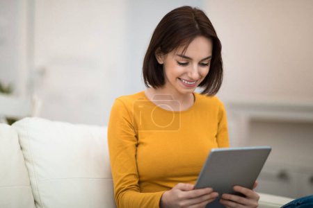 Photo for Happy pretty brunette young woman in casual outfit sitting alone on couch at home, using brand new digital pad, checking social media, chatting with friends, reading blog, copy space - Royalty Free Image