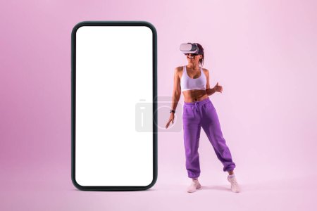 Foto de Mujer negra bailando en auriculares de realidad virtual cerca de un smartphone gigante con pantalla vacía, fondo rosa, maqueta, pancarta. dama afroamericana divertirse, explorar la tecnología 3D - Imagen libre de derechos