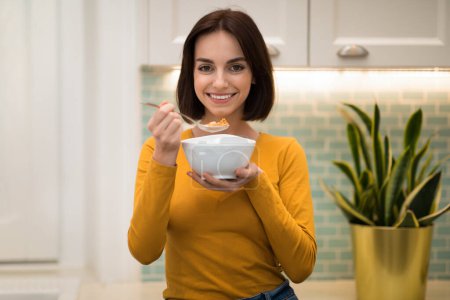 Foto de Jovencita bastante delgada alegre positiva disfrutando de un desayuno saludable en casa, mujer sonriente sosteniendo un tazón con avena, cereal o granola y cuchara, interior de la cocina, espacio para copiar - Imagen libre de derechos