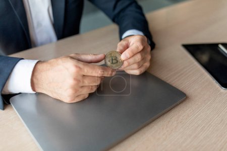 Empresario en traje sosteniendo bitcoin en sus manos, sentado en el escritorio con el ordenador portátil en el interior de la oficina, primer plano. Negocios y ahorros exitosos, dinero moderno, criptomonedas