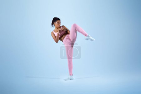 Téléchargez les photos : Entraîneur féminin noir montrant exercice de coup de pied pour la légitime défense par les jambes sur fond de studio bleu, espace de copie. Formation de femme afro-américaine en vêtements de sport en studio - en image libre de droit