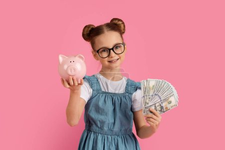 Photo for Money Saving. Cute Little Girl In Eyeglasses Holding Piggybank And Dollar Cash In Hands, Happy Female Child Enjoying Economy, Standing Isolated Over Pink background In Studio, Copy Space - Royalty Free Image