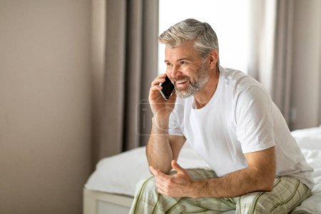 Foto de Feliz sonrisa alegre relajado atractivo de mediana edad gris de pelo hombre sentado en la cama en casa, tener conversación telefónica, mirando el espacio de copia para la publicidad, usando pijamas - Imagen libre de derechos