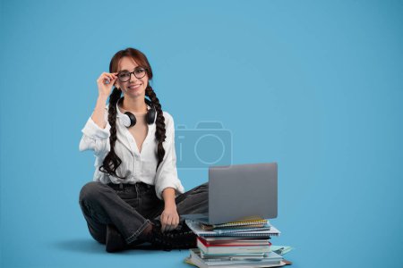 Téléchargez les photos : Glad teen girl européenne avec des nattes dans des lunettes se trouve sur le sol avec des livres, ordinateur regarde l'espace de copie isolé sur fond bleu. Travail à domicile, préparation au test, dispositif pour l'étude et l'éducation, leçon - en image libre de droit