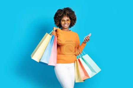Photo for Attractive cheerful millennial black woman wearing casual outfit and sunglasses shopping on smartphone, holding colorful paper bags purchases, blue studio background, sale season, black friday - Royalty Free Image