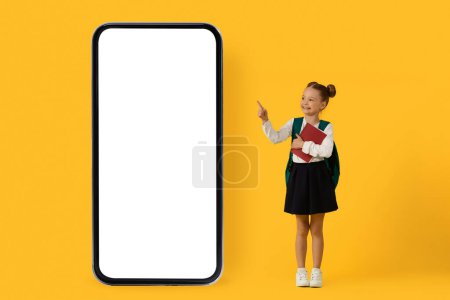 Photo for Cute Little Schoolgirl Pointing At Huge Blank Smartphone With Empty White Screen, Preteen Female Child Holding Workbooks And Showing Copy Space For Educational App Or Website, Collage, Mockup - Royalty Free Image