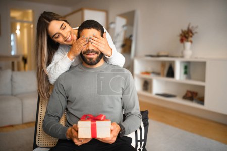 Foto de Feliz milenaria europea cierra los ojos al chico árabe, le da caja con regalo, celebrando las vacaciones en la habitación. Sorpresa para cumpleaños, aniversario y día de San Valentín, amor, relaciones en casa - Imagen libre de derechos