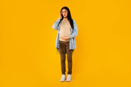 Photo for Sick pregnant woman suffering from headache, standing isolated over yellow background, full length shot. Upset expectant lady touching temples, having migraine or high blood pressure - Royalty Free Image