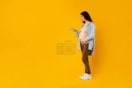 Photo for Pregnant woman looking at free space, holding open palm while standing isolated over yellow background, full length, copy space. Cheerful expectant female demonstrating place for design - Royalty Free Image