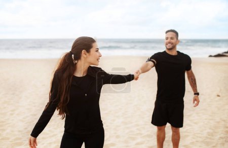 Photo for Young sporty man and woman running together on sand by sea edge, holding hands, lady looking back at her husband, enjoying workout on nature - Royalty Free Image