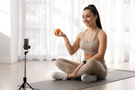 Photo for Cheerful beautiful brunette sporty young woman famous fitness blogger influencer streaming from home, sitting on yoga mat with tape measure on her neck, showing orange and smiling, using smartphone - Royalty Free Image