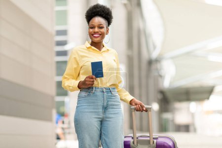 Foto de Billetes de viaje baratos. Mujer negra feliz con maleta sosteniendo el pasaporte sonriendo a la cámara de pie en el aeropuerto al aire libre. Una foto de la dama turista esperando el vuelo. Concepto de oferta de vacaciones y turismo - Imagen libre de derechos