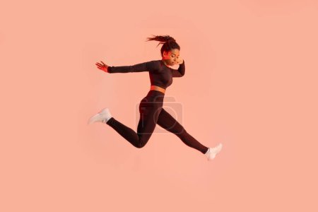 Téléchargez les photos : Studio plan de jeune femme noire en forme sautant sur fond de studio lumière au néon pêche, espace libre. Femme afro-américaine en vêtements de sport faisant de l'exercice et sautant dans l'air - en image libre de droit