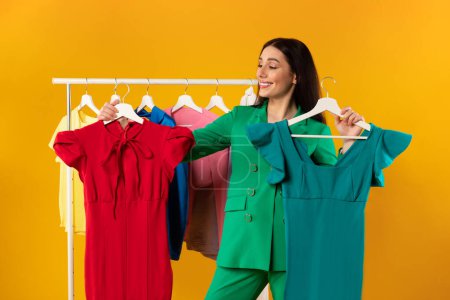 Photo for Happy shopaholic woman choosing between two new dresses, standing near clothing rail over yellow studio background. Fashion and trends concept - Royalty Free Image