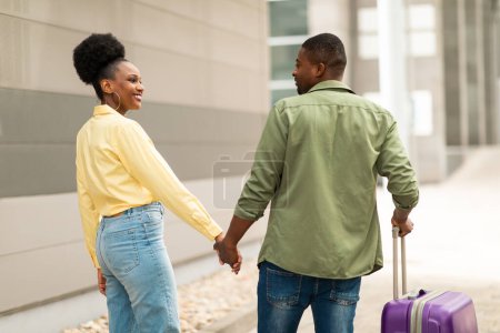 Téléchargez les photos : Des vacances. Heureux conjoints afro-américains voyageant à pied avec valise tenant la main posant à l'aéroport en plein air. Transport et Tour Offre Concept. Vue arrière prise - en image libre de droit