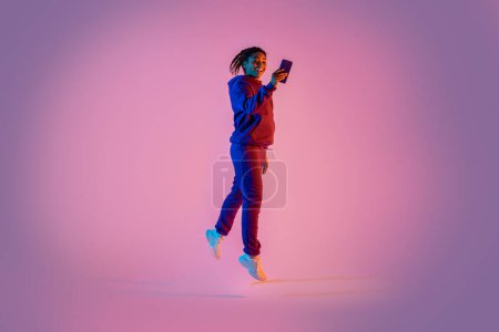 Foto de Longitud de cuerpo completo de la joven mujer negra deportiva en ropa deportiva saltando y utilizando el teléfono móvil sobre fondo de neón rosa. Concepto deportivo de entrenamiento - Imagen libre de derechos