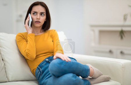 Photo for Annoyed beautiful brunette young woman in comfy casual outwear sitting on couch with legs up, having phone conversation with friend or lover at home, have fight, looking aside, copy space - Royalty Free Image