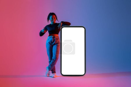 Foto de Mujer negra feliz de pie cerca de un gran teléfono inteligente con pantalla en blanco y apuntando a la pantalla, de pie en la luz de neón, maqueta para la aplicación móvil, sitio web, el diseño de su anuncio - Imagen libre de derechos