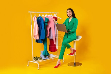 Photo for Happy shopaholic lady shopping online via laptop computer, sitting near clothing rack in studio on yellow background, full length. Fashion and trends concept - Royalty Free Image