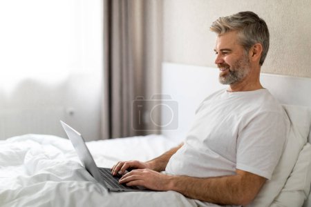 Téléchargez les photos : Relaxé heureux bel homme d'âge moyen portant un pyjama en utilisant un ordinateur portable PC moderne au lit, surfer sur Internet, vérifier les médias sociaux, lire les nouvelles ou travailler en ligne, copier l'espace, vue de côté - en image libre de droit