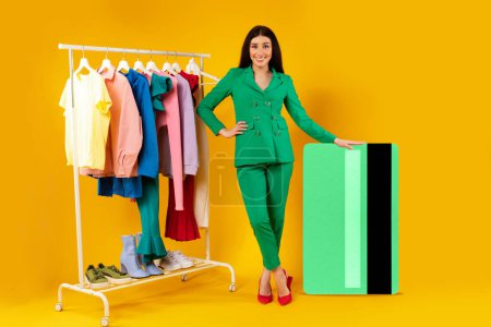 Photo for Mobile shopping. Happy lady customer standing near huge credit card and clothing rail over yellow background, full length. Contented shopaholic buying clothes online - Royalty Free Image