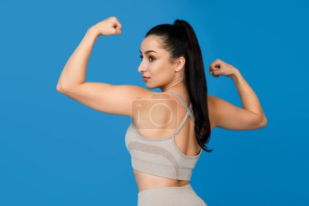 Téléchargez les photos : Jolie jeune femme sportive brune en tenue de sport bodybuilder féminin montrant ses muscles des bras, démontrant biceps musculaires, triceps, dos sur fond de studio bleu - en image libre de droit