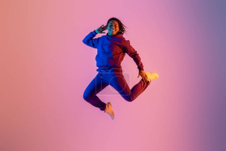 Téléchargez les photos : Joyeuse femme noire énergique en vêtements de sport s'amuser et sauter haut sur fond de studio coloré néon, pleine longueur, espace libre, bannière - en image libre de droit