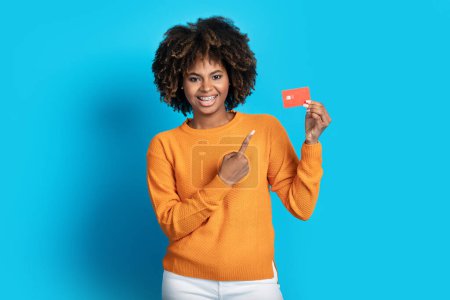 Photo for Happy smiling pretty millennial african american woman in casual pointing at red bank card in her hand, recommending easy online banking, blue studio background - Royalty Free Image