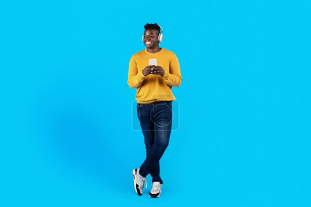 Téléchargez les photos : Souriant Black Guy dans les écouteurs sans fil tenant Smartphone et regardant loin tout en se tenant sur fond de studio bleu, Happy African American Man écouter de la musique en ligne sur téléphone mobile, pleine longueur - en image libre de droit