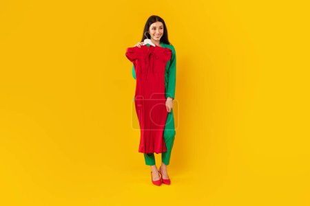 Photo pour Plan complet de la créatrice de mode femme debout avec cintre à la main montage sur robe élégante rouge, posant sur fond jaune, espace libre - image libre de droit