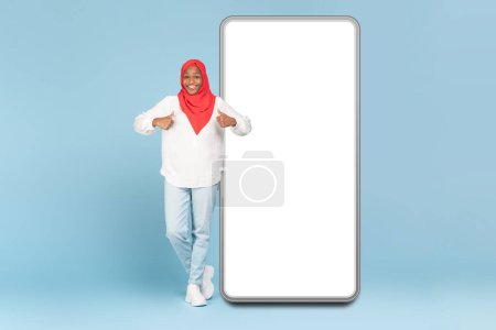 Téléchargez les photos : Femme noire joyeuse en hijab debout près d'un énorme nouveau téléphone portable avec écran blanc vide et montrant pouces levés, maquettes, recommander l'application mobile, fond bleu, pleine longueur - en image libre de droit