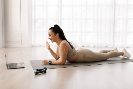 Photo for Side view of happy beautiful brunette well-fit athletic young woman thankful for online workout, lying on yoga mat, waving at laptop screen and smiling, home interior, copy space - Royalty Free Image