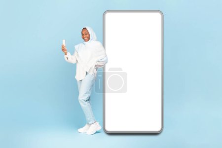 Foto de Mujer afroamericana feliz en hijab de pie cerca de un gran teléfono celular con pantalla blanca vacía y el uso de dispositivos móviles, maqueta para el diseño de sitios web o aplicaciones, fondo azul - Imagen libre de derechos