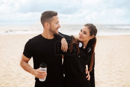 Photo for Young sporty couple in love in black sportswear embracing after warming up on sea ocean beach outdoors, standing by seaside and looking at each other - Royalty Free Image