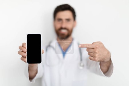 Photo for Medical Ad. Male Doctor Wearing Uniform Pointing At Blank Smartphone In His Hand While Standing Near White Wall In Hospital, Smiling Physician Man Recommending Online Offer, Selective Focus, Mockup - Royalty Free Image