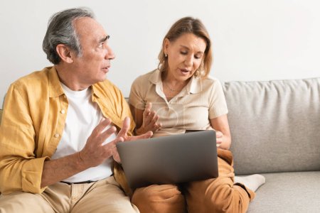 Photo for Displeased senior couple arguing, looking at their online bank account on laptop, woman using computer and shopping online at home, free space - Royalty Free Image