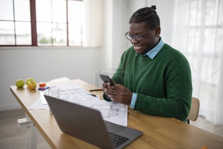 Foto de Hombre de negocios negro sonriente usando su teléfono trabajando y comunicándose en línea sentado en el ordenador portátil en la oficina moderna. Hombre Gerente de mensajes de texto y el uso de aplicaciones comerciales en el teléfono inteligente en el lugar de trabajo - Imagen libre de derechos