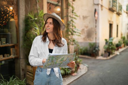 Foto de Sonriente inspirada milenaria turista dama europea en sombrero con mapa mira los lugares de interés en busca de viajes de ruta en la ciudad, al aire libre. Emociones del viaje en vacaciones, impresiones del viaje, anuncio y oferta - Imagen libre de derechos