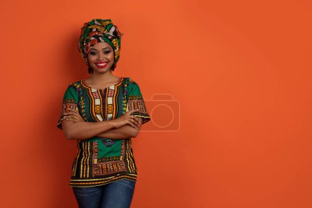 Téléchargez les photos : Positif attrayant jeune femme noire en costume africain traditionnel coloré posant avec les bras croisés sur la poitrine sur fond de studio orange, souriant à la caméra, debout par l'espace libre pour la publicité - en image libre de droit