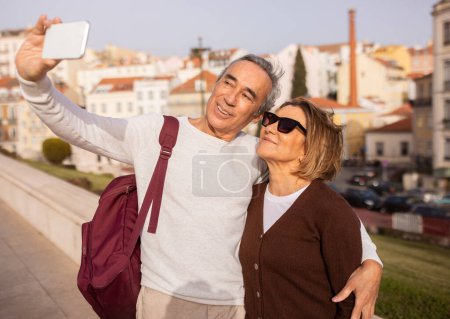 Téléchargez les photos : Voyage de vacances. Heureux couple de touristes seniors faisant Selfie sur téléphone étreinte debout avec sac à dos à l'extérieur. Époux voyageant à pied à Lisbonne Profitant d'une visite de la ville. Tourisme et visites guidées - en image libre de droit