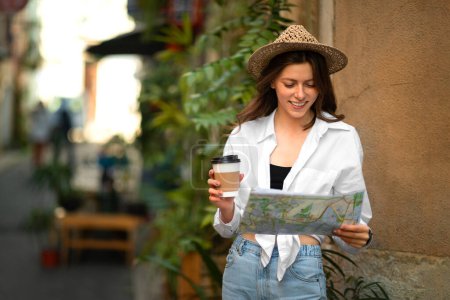 Foto de Glad millennial turista europea en sombrero con mapa en busca de viajes de ruta y hotel en la ciudad, disfrutar de una taza de café para llevar, al aire libre. Viajar de vacaciones, descansar, viajar en tiempo libre - Imagen libre de derechos