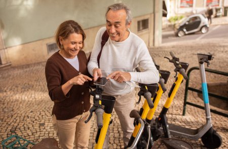 Téléchargez les photos : Heureux couple d'âge mûr en utilisant Smartphone Location Scooters électriques à l'extérieur. Personnes âgées voyageant dans une ville européenne Profitant de vacances ensemble. Tourisme et vie active à la retraite - en image libre de droit