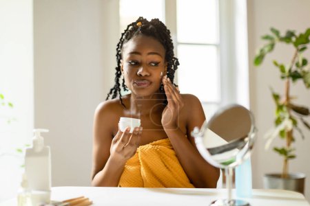 Téléchargez les photos : Belle femme noire appliquant de la crème pour le visage et regardant miroir, peau hydratante avec lifting crème de jour nourrissante à l'intérieur de la chambre, espace libre - en image libre de droit